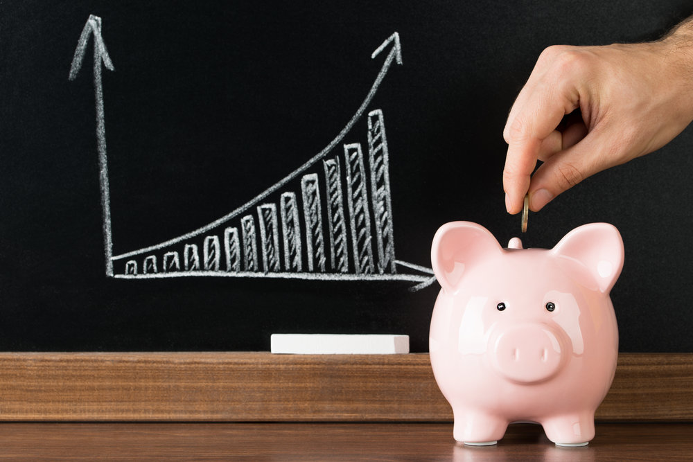 Hand Putting A Coin Into Piggy Bank In Front Of Blackboard Showing Graph