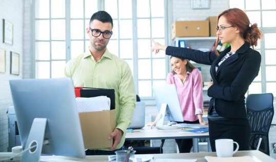 Redhead businesswoman pointing at the door to dismissed employee. Smiling female coworker behind. All with formalwear. Man holding a box with documents.