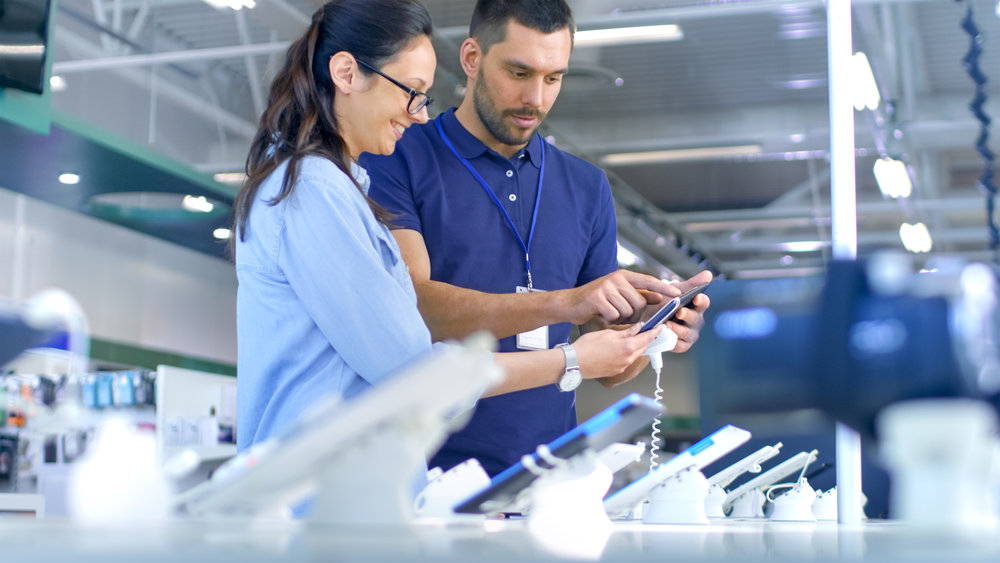 In the Electronics Store Consultant Gives Professional Advice to a Young Woman, She Considers Buying New Tablet Computer and Needs Expert Opinion. Store is Modern, Bright and Has all the New Devices.