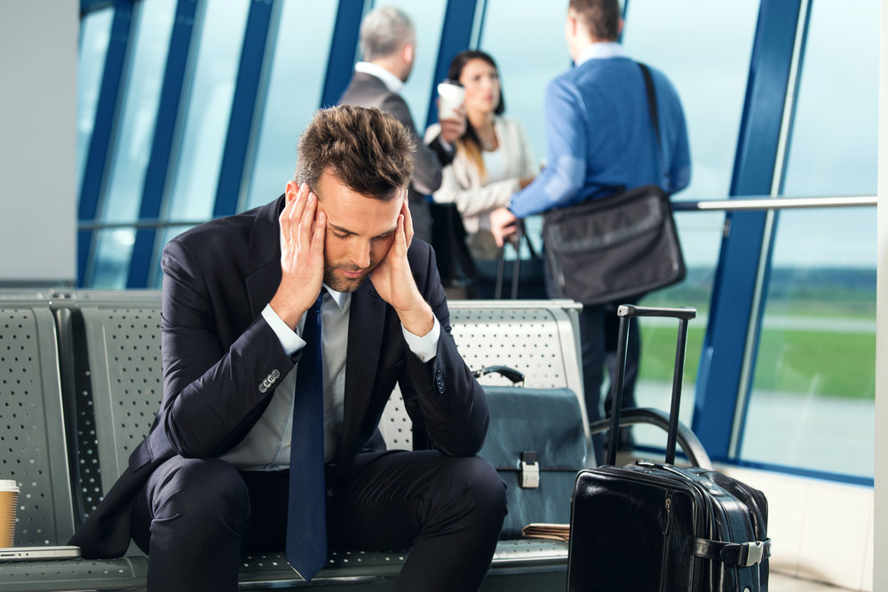 Businessman waiting for a flight at the airport lounge, sitting tired on a banch, group of people standing in the background.