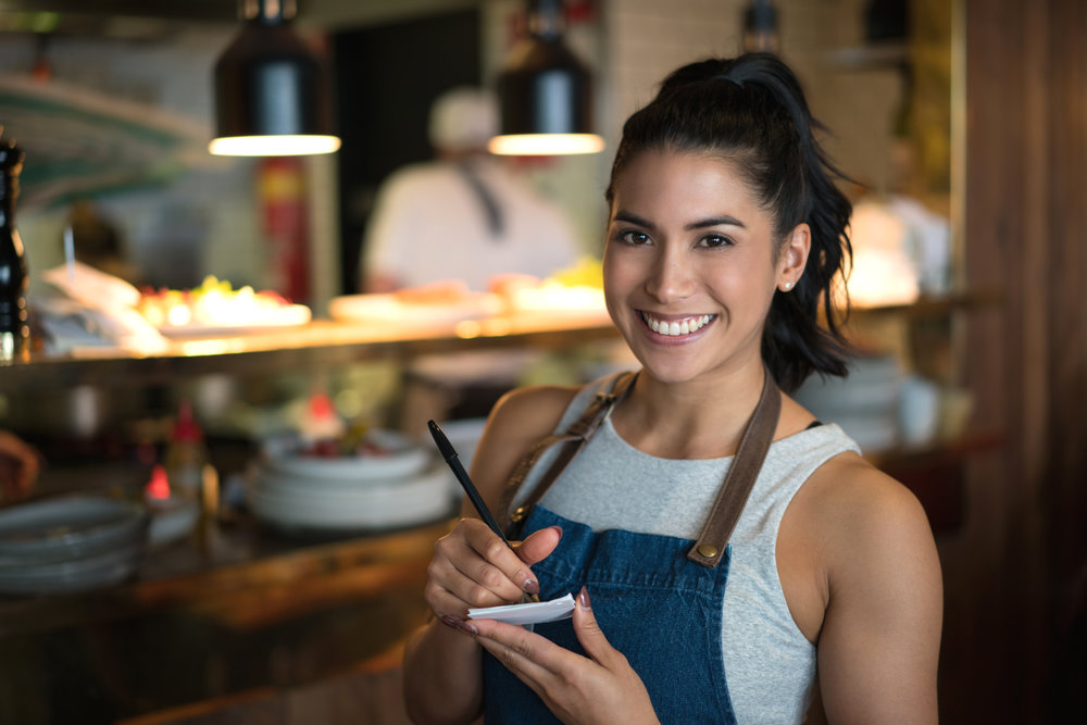 Happy waitress working at a coffee shop and looking at the camera holding a notepad