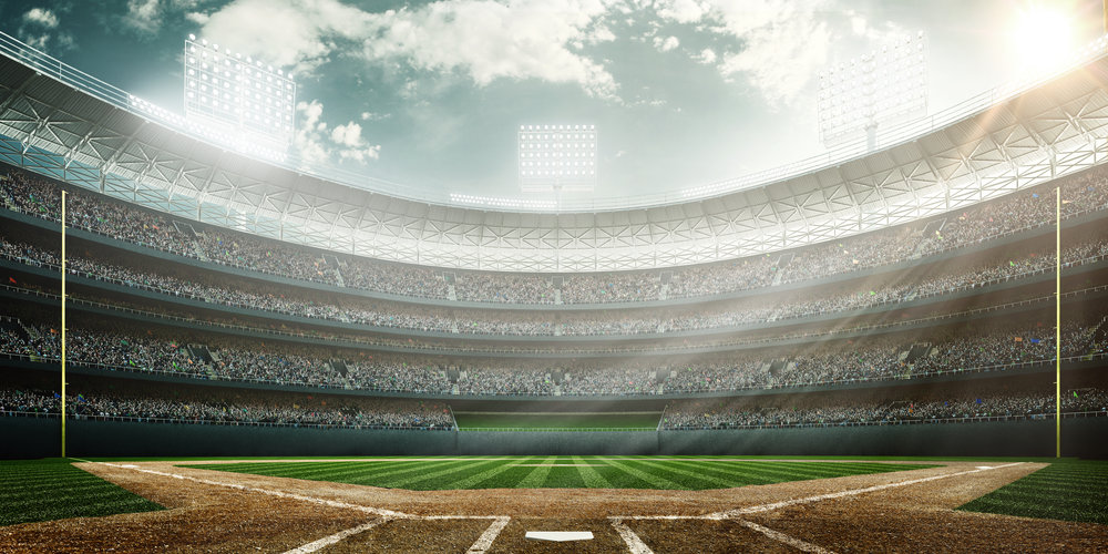 A wide angle of a outdoor baseball stadium full of spectators under a cloudy sky at midday. The image has depth of field with the focus on the foreground part of the pitch.