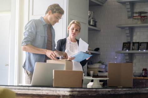 A photo of woman with clipboard watching man wrapping presents. Young couple preparing list of gifts for Christmas. Both are in casuals in domestic room.