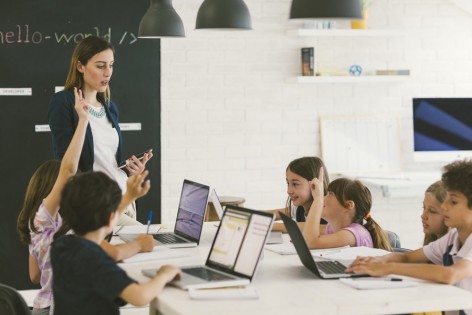 Group of children at coding class. They are sitting by the desk and using laptops. Their female teacher standing beside desk, holding digital tablet and asking question and children raising hands to answer.