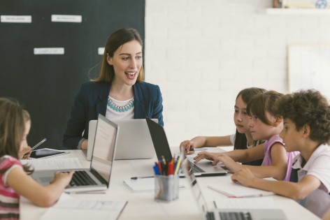 Group of children at coding class. Children and their teacher sitting by the desk and using laptops. Teacher talking to kids.