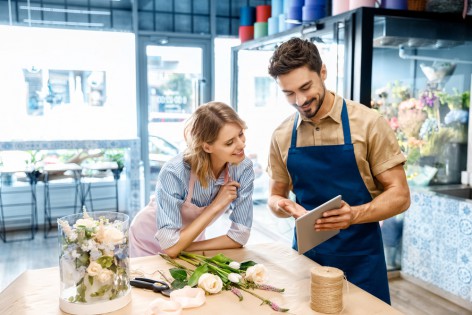 smiling young florists in aprons using digital tablet while working together in flower shop