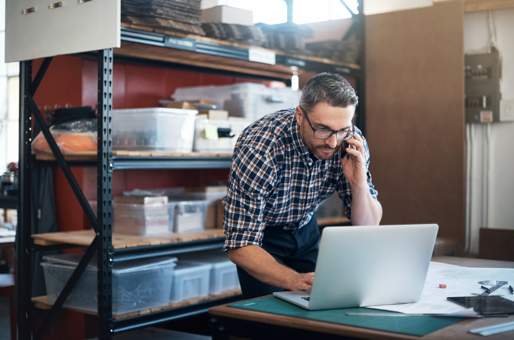 Shot of a man using a laptop and mobile phone while working on a project in a workshop